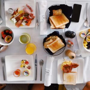 Flatlay of an elegant brunch setup with fruits, toasts, and beverages on a white tablecloth.