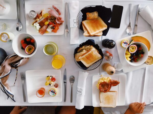 Flatlay of an elegant brunch setup with fruits, toasts, and beverages on a white tablecloth.