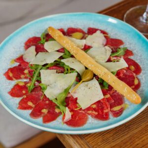 Close-up of carpaccio served with arugula, cheese, and a breadstick on a blue plate.