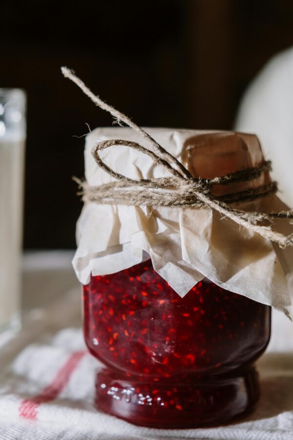 A close-up of homemade raspberry jam in a rustic glass jar on a white table.