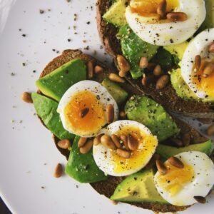 Delicious avocado toast topped with soft-boiled eggs and pine nuts on a white plate.