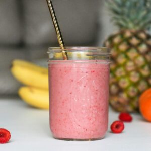 A refreshing pink smoothie made with bananas, raspberries, and tropical fruits in a glass jar.